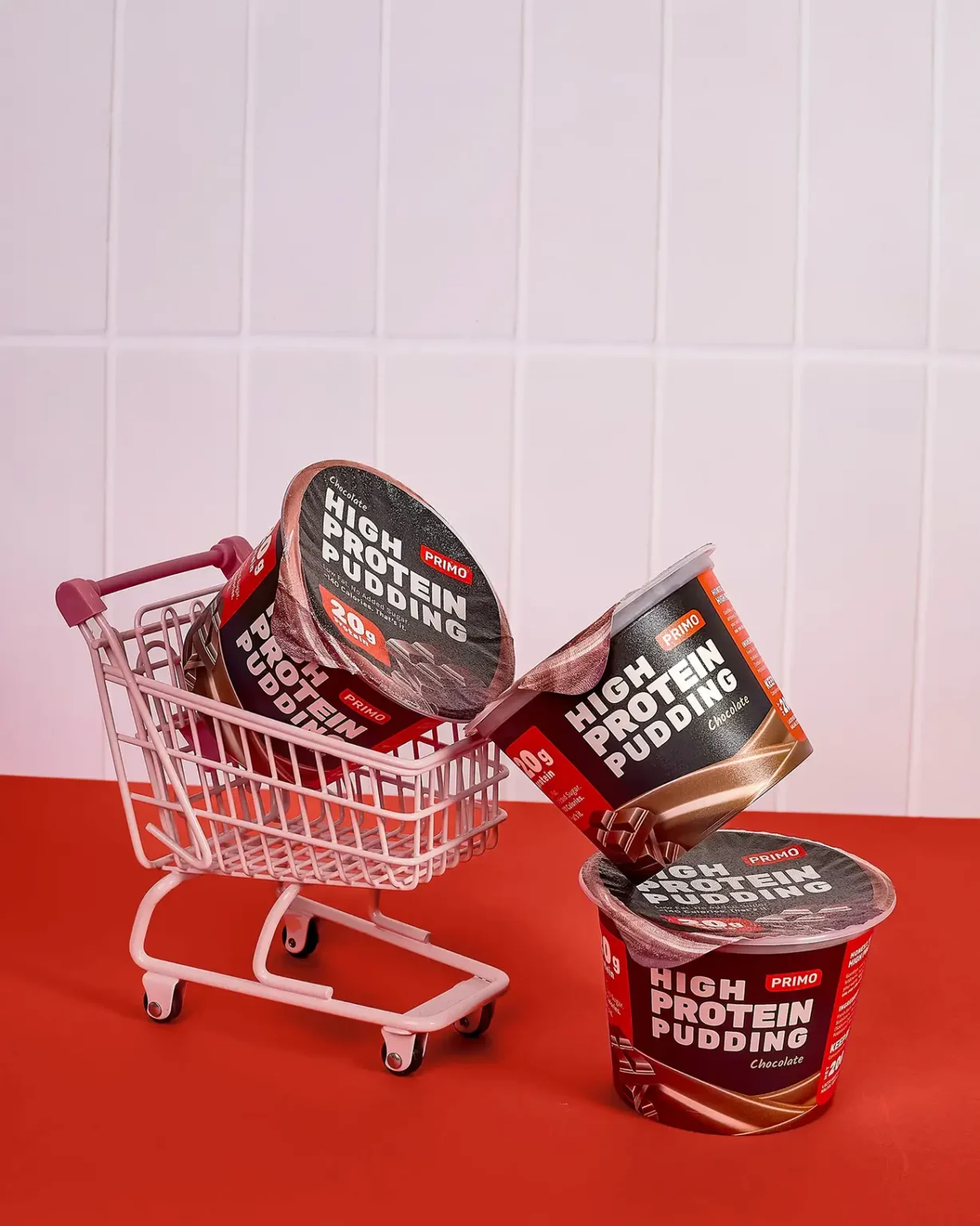 Three Primo Chocolate High Protein Pudding cups in and around a playful mini shopping cart on a red surface with a tiled wall background.