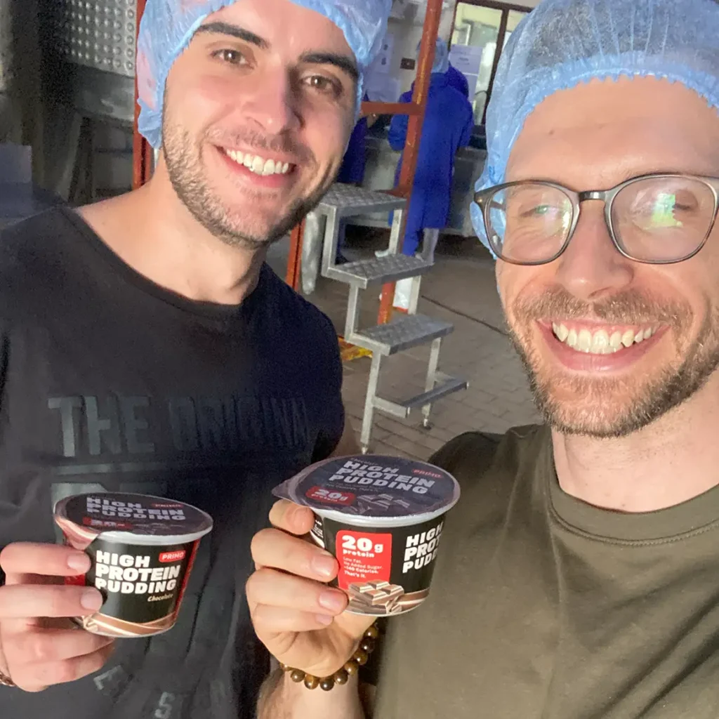 Two Primo founders smiling in hairnets and holding the very first cups of Primo Chocolate High Protein Pudding fresh from production.