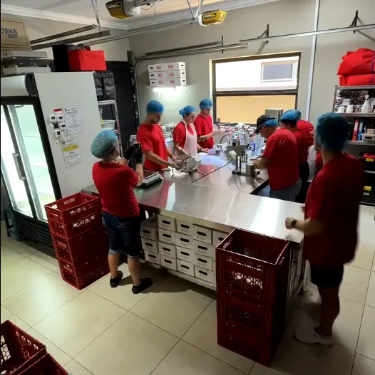 Primo team in red shirts and blue hairnets working in the small garage factory to hand-pack high protein puddings.